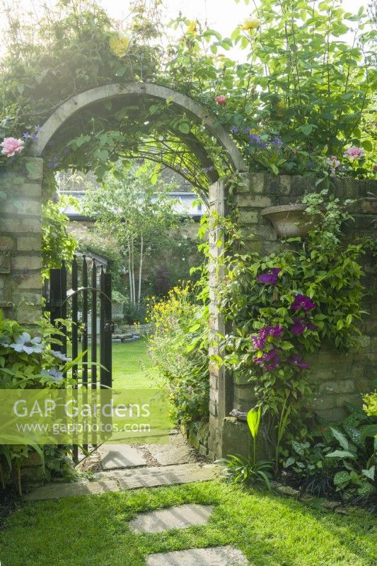 Wrought iron garden gate in a brick wall clothed with roses and clematis. Stepping stones leading across a neatly mown lawn. June