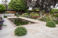 Brick edged rectangular pool on terrace of sandstone paving with garden furniture.  Border plants include David Austin roses; Salvia nemorosa 'Caradonna', Lavender, Ballota and Pittosporum topiary balls