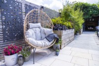 Wicker egg chair on white tile patio next to wooden raised bed planted  with Heuchera, Ferns and Japanese maple with decorative panels on fence and potted Fuchsia, Thyme, Alchemilla and sempervivum on the ground