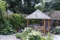 Round wooden gazebo with tile roof surrounded by mixed planting in garden borders
