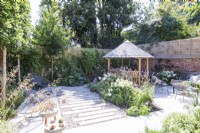 Garden with stone tile patio and staggered path with mixed planting beds and borders, seating areas and a round wooden gazebo with a tile roof