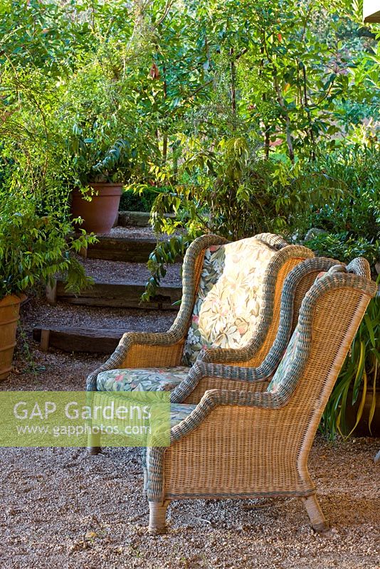 Two wicker arm chairs in front of a gravel step with plants in clay containers and backed by high-growing plants