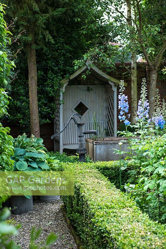 A shady corner of a walled garden with covered wooden seat, a small pond made from a galvanised metal animal feeding trough and shade tolerant planting including Hosta and box hedges.