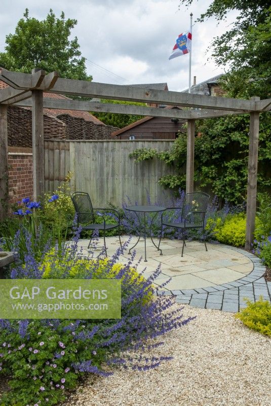 Gravel path leading to circular area of slabs and setts under pergola frame with metal table and chairs and bordered by shrubs and perennial plants including Nepetia - Catmint, Irises and Hardy Geraniums  -  Hidden Gardens Day, Woodbridge, Suffolk