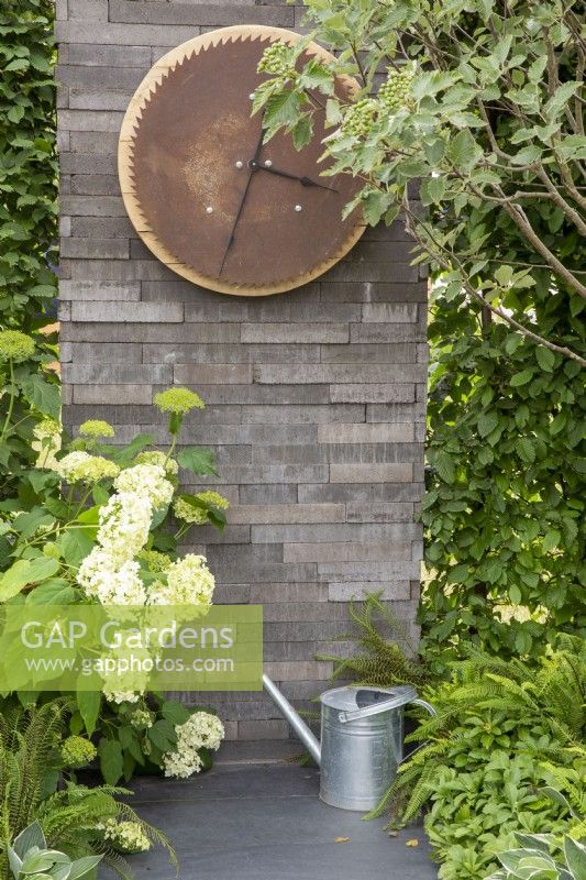 Wall made from stacked concrete slabs, outdoor clock using reclaimed materials, stone paving with metal watering can and Hydrangea arborescens 'Annabelle'