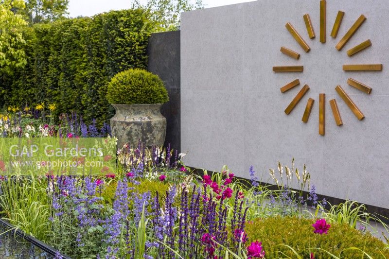 A colourful perennial bed planted with Nepeta, Salvia and Ixia 'Mabel'. A giant, modern copper clock decorates a tiled wall. A large ceramic vase with a yew ball and yew hedge in the background. June. Designer: Kevin Dennis, Bord Bia Bloom 2023