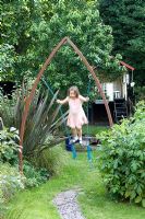 Girl playing in town garden on swing hanging from metal arch. Phormium on left hand side and wooden treehouse behind. Yulia Badian garden, London, UK