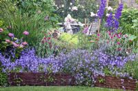 Raised bed with woven willow edging - Lychnis coronaria, Campanula poscharskyana, Delphinium and Hydrangea. Seating area with table and chairs beyond - Scheper Town Garden 
