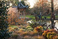 Evening view across the lake at Seven Acres to the Chinese pagoda with Hamamelis Aphrodite, Carex Flagellifera and Carex Ashimensis evergold, winter. RHS Garden, Wisley.  