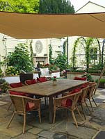 Walled courtyard with dining table shaded by suspended sail. Raised beds planted with figs, hydrangea, begonia, petunia. Reclaimed brick and stone. Metal pergola with wisteria.