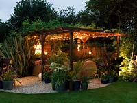 Night view of pergola seating area, flanked by phormium. Vine on pergola supporting hanging lanterns and rope lights. Pots of agapanthus, bamboo, acer, fern, canna, palm, geranium.