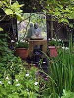 Hidden in shady corner beneath magnolia tree, a tiny water feature with statue of Buddha perched above miniature waterfall, a mirror behind. Edged in white iris, geranium.