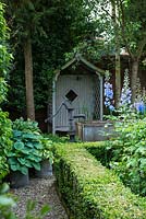 A shady corner of a walled garden with covered wooden seat, a small pond made from a galvanised metal animal feeding trough and shade tolerant planting including Hosta and box hedges.