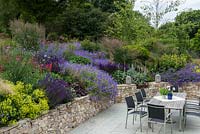 A patio dining terrace alongside a large bank planted with perennials and ornamental grasses. Planting includes: Salvia 'Caradonna', Geranium 'Rozanne', Campanula persicifolia, Alchemilla mollis, Penstemon 'Garbet' and Molinia 'Karl Foerster'.