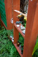 Wooden garden divider with niches for ornaments and found items including a tin aeroplane model, bottles and pebbles. Green foliage planting. 