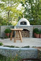 Outdoor pizza oven surrounded by pots of herbs,  with table and Cow Parsley in a jug - The Refuge Garden in aid of Help Refugees UK, RHS Malvern Spring Festival 2017 - Design: Sue Jollans, Sponsors: Readyhedge, Everedge, Outchester and Ross Farm Cottages