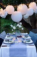 Outdoor dining table dressed in shades of blue and with a vase of white Japanese anemones.  Paper ball lanterns, fairy lights and paper pompoms hang over the table to decorate. Pockets are made in the folded napkins to put name card and each setting is finished with an olive branch.  Tealights are put on the table to light it up as dusk falls