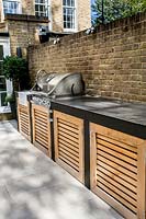 Contemporary garden with white stone patio, with a garden kitchen
 built in barbecue against brick wall.