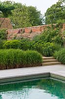 Edge of swimming pool with paved poolside and steps up to a walled garden with borders of ornamental grasses and perennial flowers
