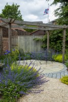 Gravel path leading to circular area of slabs and setts under pergola frame with metal table and chairs and bordered by shrubs and perennial plants including Nepetia - Catmint, Irises and Hardy Geraniums  -  Hidden Gardens Day, Woodbridge, Suffolk