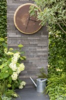Wall made from stacked concrete slabs, outdoor clock using reclaimed materials, stone paving with metal watering can and Hydrangea arborescens 'Annabelle'