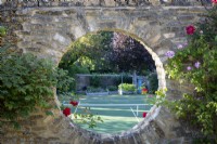 A moon window in a stone wall overlooking a tennis court, framed with Rosa 'Ferdinand Pichard'