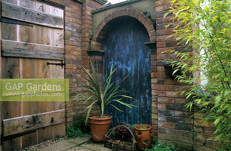 Corner of patio with copper clad alcove and rustic oak door. Cordyline in container. 