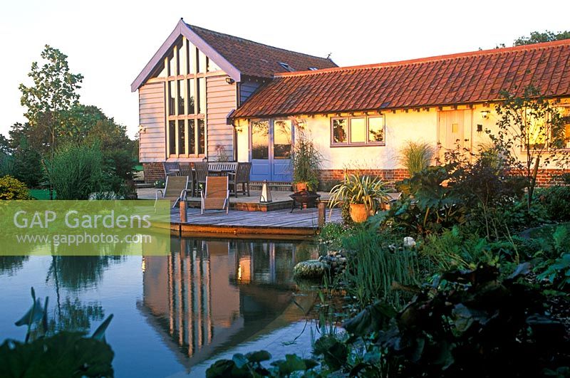 View across swimming pond to timber decking, outside eating area with table and chairs and converted barn with warm autumn sunlight reflected on building