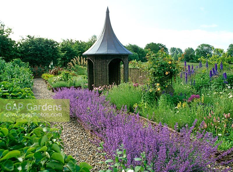 Willow summerhouse with lead covered roof in cottage garden with willow fences and raised beds, Nepeta 'Walker's Low', Delphiums, roses, Persicaria bistorta 'Superba' and Alliums