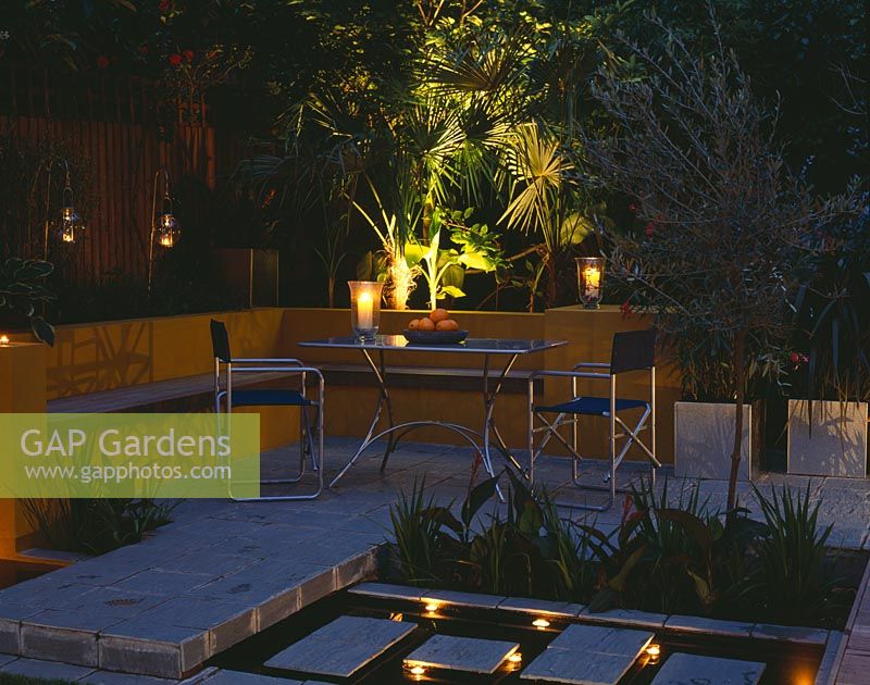 Aluminium table and chairs on patio surrounded by yellow rendered walls with raised beds and rill. Evening lighting with candles and lanterns. 