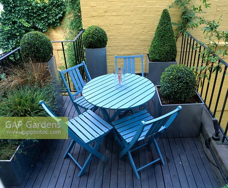 Roof garden with decked terrace and blue table and chairs, yellow painted wall and metal containers planted with Buxus - Box topiary