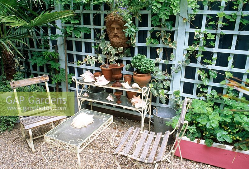 Seats in shady gravel area beside metal and glass etagere and table - antique terracotta mask mounted on trellis