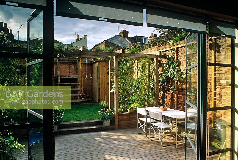 Small urban courtyard garden with pergola, lawn, childs play area and dining area viewed from inside house - Acton, London

  