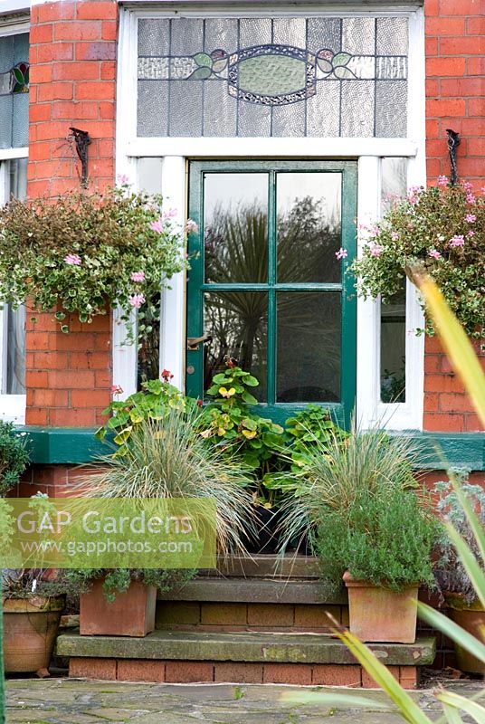 Green back door to a traditional Victorian red brick house with stained glass surround. Raised steps with pots of geranium, grasses and herbs