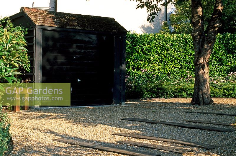 Concrete garage painted black to imitate black timber in urban front garden with gravel and timber driveway. Existing Cherry tree and Laurel hedge.
