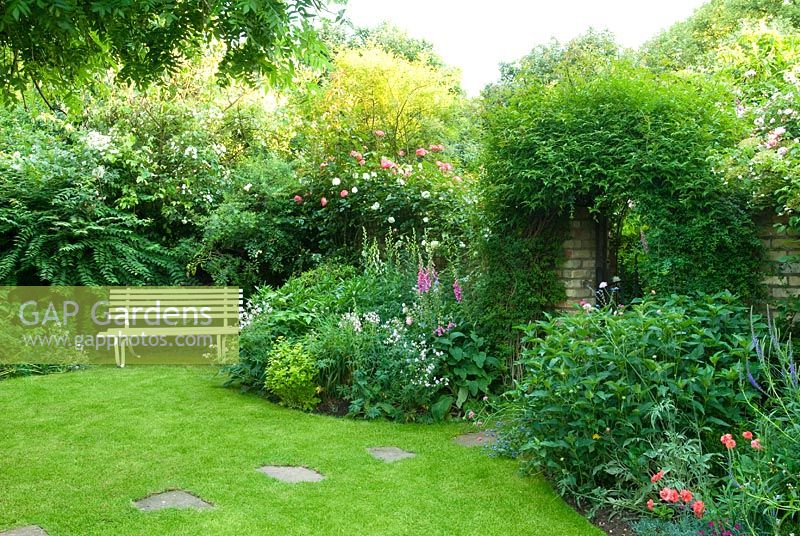 Lawn with stepping stones leading to gate in garden wall, herbaceous border with roses and jasmine and white garden bench - New Square, Cambridge