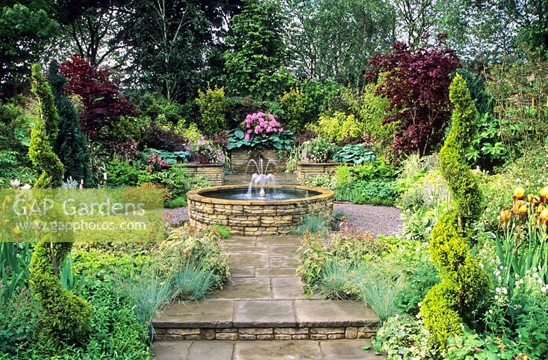 Formal water garden with raised circular pond and fountain on patio on sloping site with mixed borders and sprial topiary