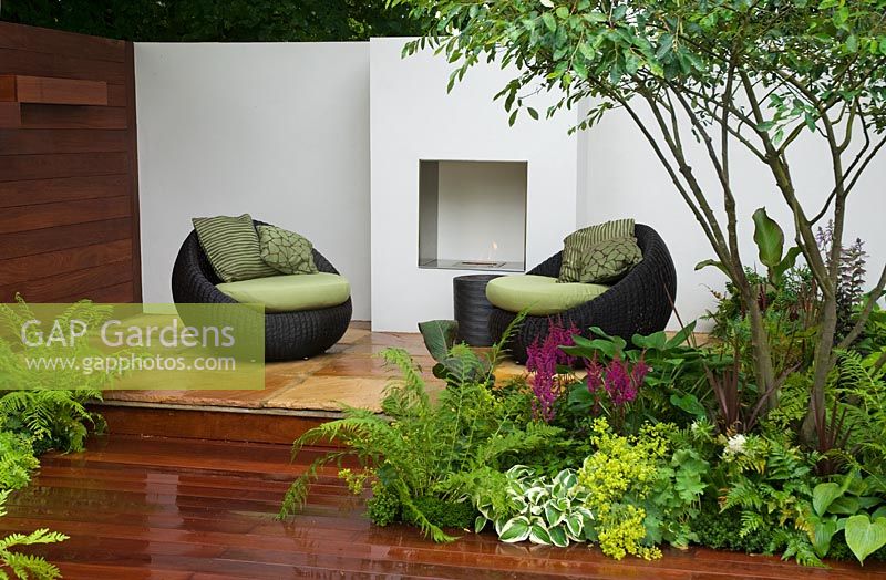 Round wicker chairs with green cushions in front of outdoor fireplace with ferns and Astilbe in the foreground in the Formal Elements Garden - RHS Hampton Court Flower Show 2008