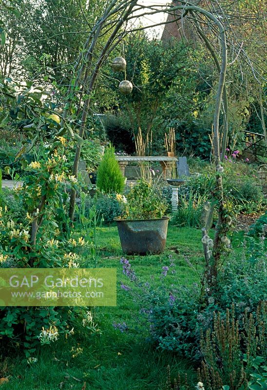 Looking through a Hazel arch with Lonicera japonica 'Halliana' to a 'secret garden' with recycled copper planter