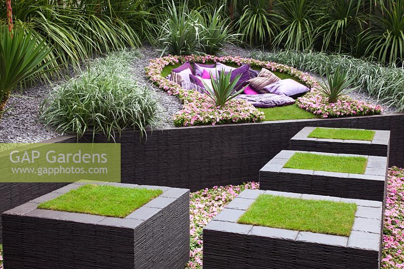 Raised, turf covered slate stepping stones lead through a sunken bed of Impatiens 'Cupido Pink' towards a seating area surrounded by sharp and spiky planting of Cordyline australis, Agave americana, Yucca gloriosa, Yucca recuvifolia, Yucca floribunda, Chamaerops humilis cerifera and Astelia chathamica 'Silver Spear' in the The Quilted Velvet Garden, sponsored by Quilted Velvet and Hortus Infinitus - Silver Flora medal winner at RHS Chelsea Flower Show 2009