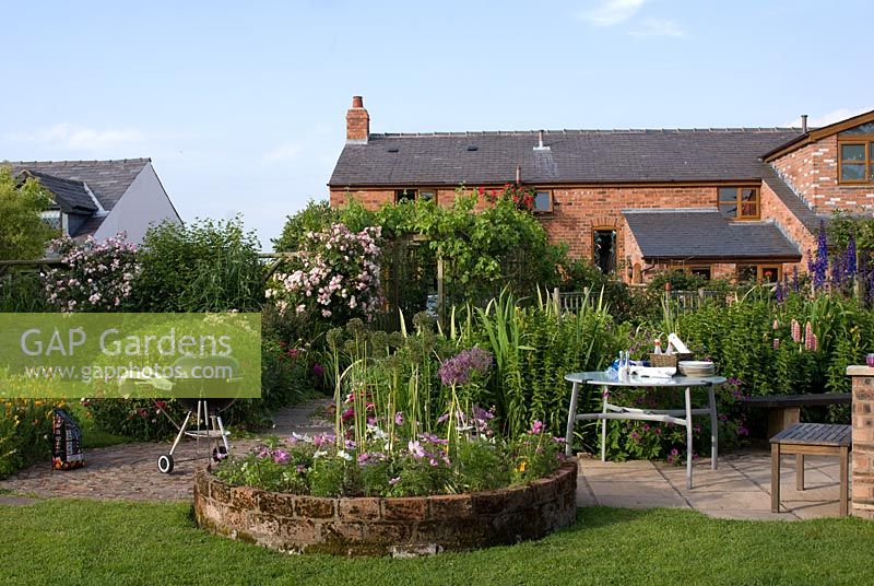 Barbeque and eating area on patio in country garden with adjacent herbaceous borders, trellis with climbing roses and a circular bed with Cosmos and Allium 