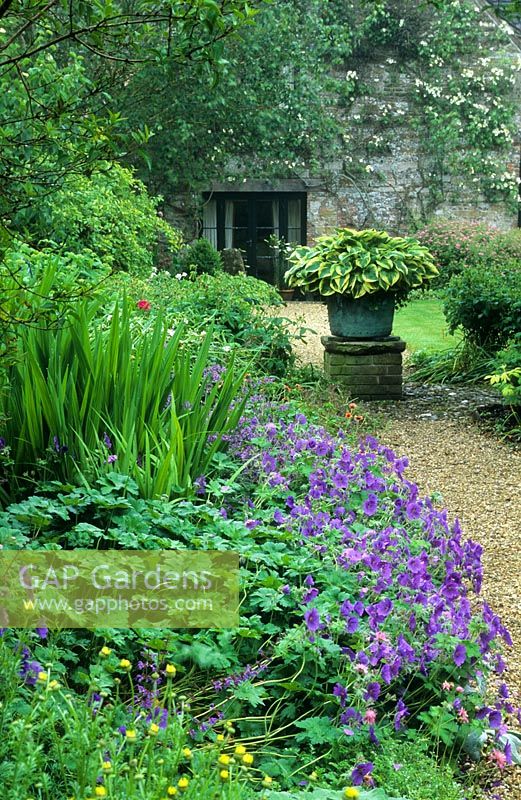 View to house, flint cobble circle and old 'copper' with variegated Hosta on a raised plinth. Mixed border with Geranium x magnificum and Crocosmia foliage - Cerne Abbas, Dorset