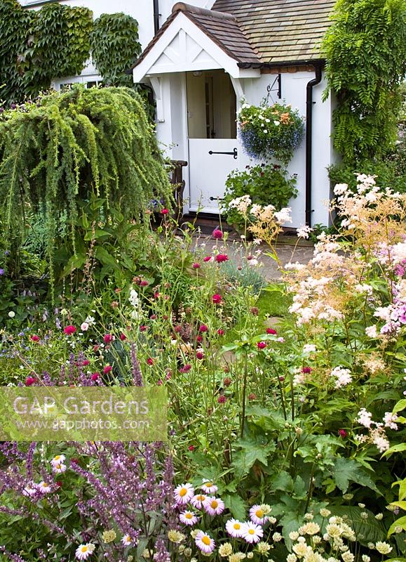 Borders of herbaceous perennials black and white barn door with well planted hanging basket with flowers in profusion, packed into an idyllic English cottage garden, at Grafton Cottage, NGS , Barton-under-Needwood Staffordshire