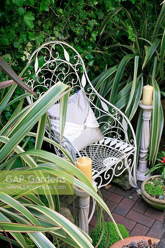 Ornate white wirework chair flanked by variegated Phormiums and candlesticks makes a striking focal point in a secluded town garden