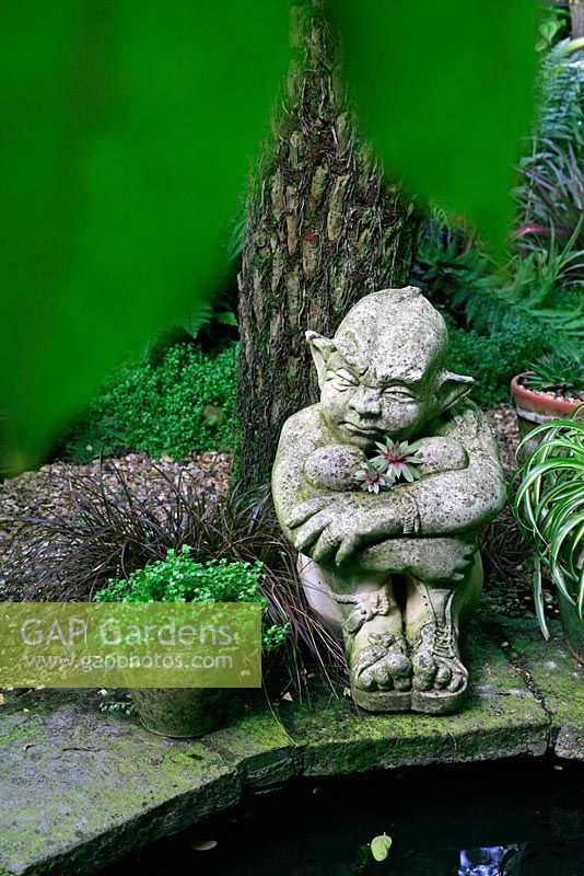 Sculpture of an imp sitting beneath the trunk of a tree fern beside a shallow pond with houseleeks, Sempervivum sprouting from his body