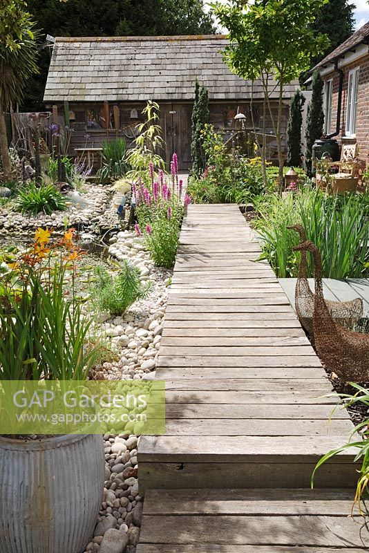 Seaside Inspired garden. Timber decking pathway through seaside garden leading to rustic beach hut. Crocosmia planted in old metal dolly tub, wirework geese sculptures, Lysimachia - Loosestrife and pebble beach.