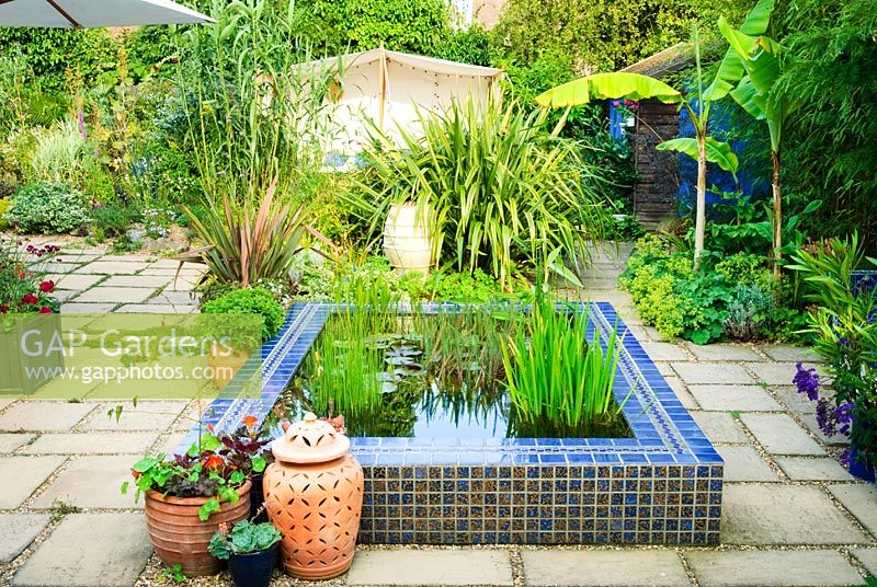 Paved courtyard with raised pool clad in blue glazed tiles. View to Moroccan style outdoor tent. Phormiums, Musa 'Basjoo', Bamboo, Arundo donax in borders. Decorative terracotta planters and containers