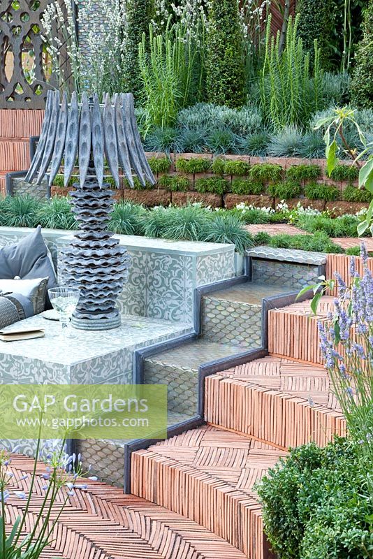 Tiled outdoor seating area with wooden lamps surrounded by a herringbone path, plants include Lavandula angustifolia 'Alba', Koeleria glauca and Armeria maritima 'Alba'. 'The Garden Lounge' - Silver Gilt Medal Winner - RHS Hampton Court Flower Show 2010 