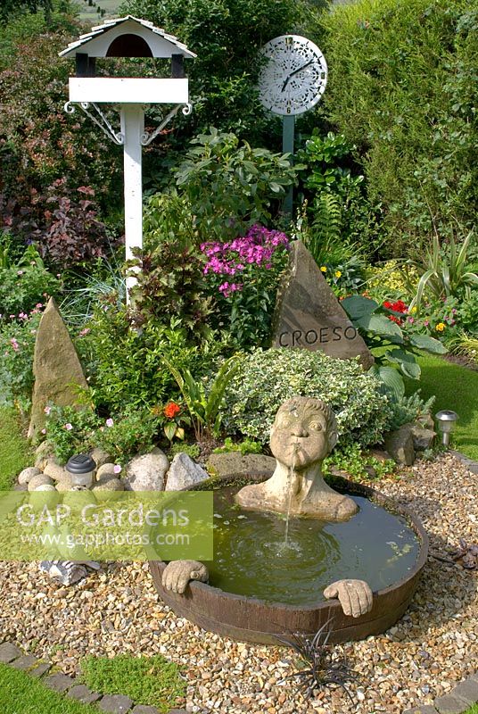 Corner of small back garden with mixed borders, Leylandii hedge and features including - a painted bird table, a half barrel water feature with submerged head spout and hands, clock face made from painted ornate table top and carved stone sculpture with 'Croeso' - welcome in Welsh at 'Trevinia', Stubbins, Lancashire NGS