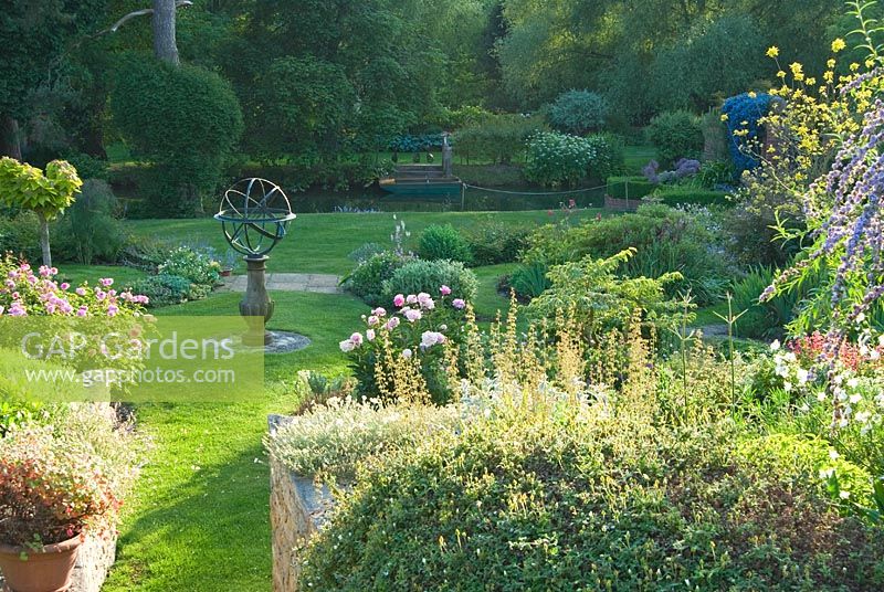 The upper garden has a sundial at its centre, and shaped beds full of herbs, herbaceous perennials including Peonia and interesting shrubs with mill pond seen beyond. Mill House, Netherbury, Dorset, UK
